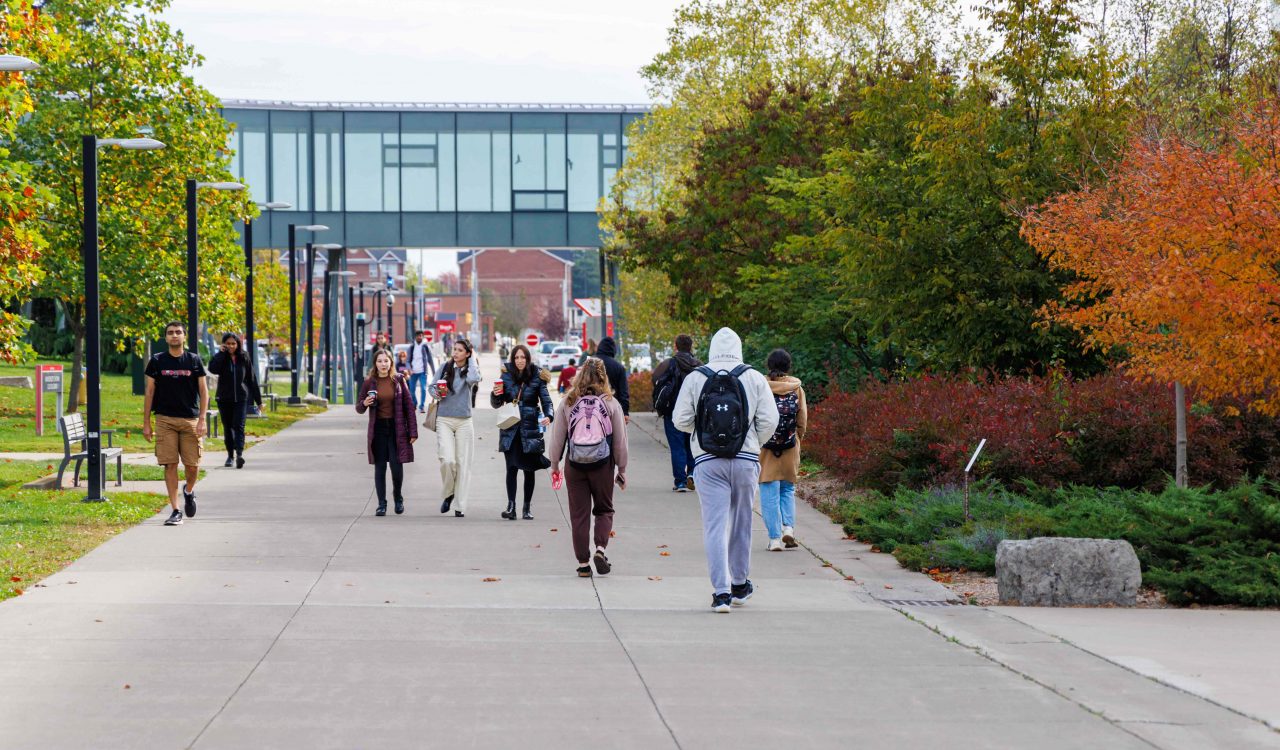 Students walk along an outdoor concrete concourse with a glass walkway overtop.