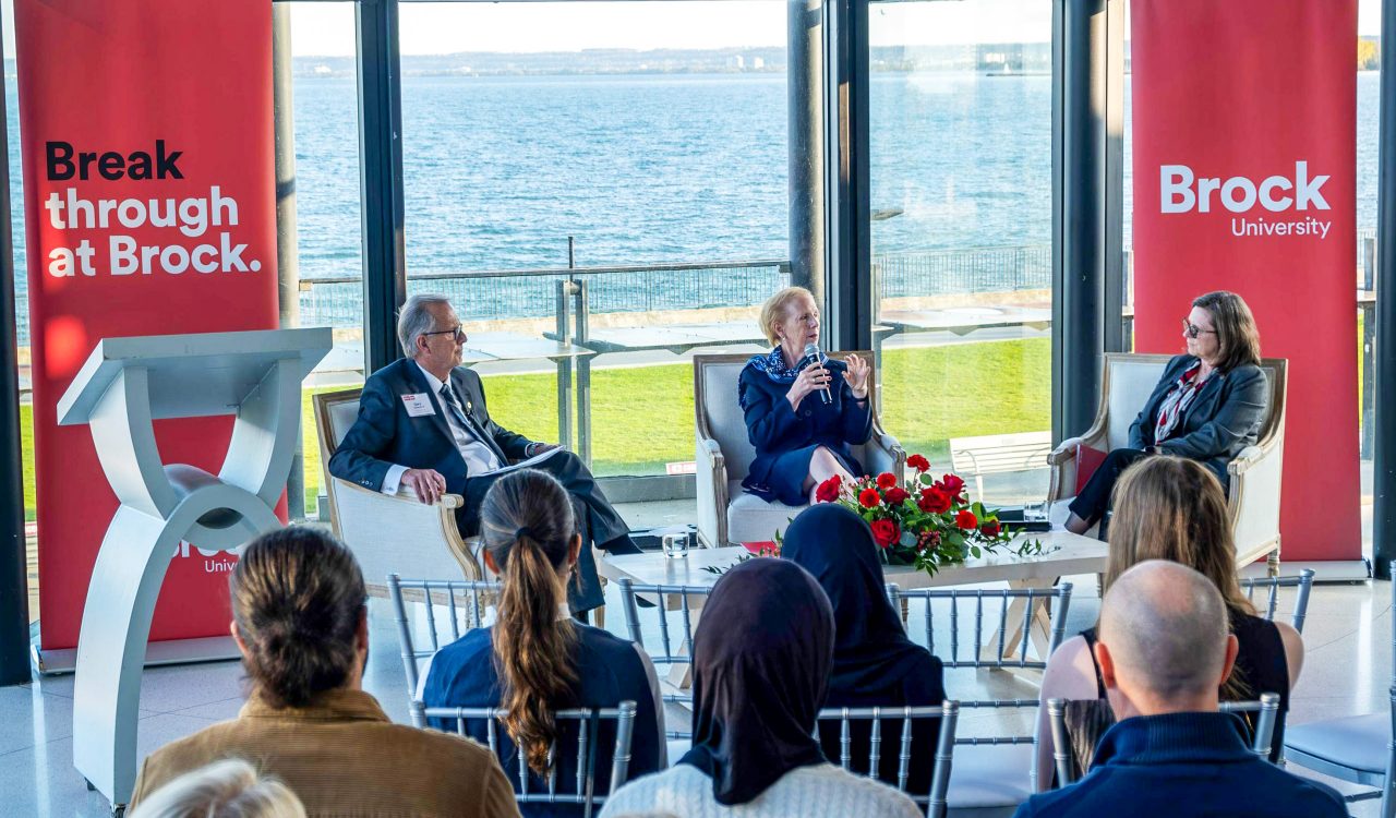 Three people sit at the front of an audience in a sunny room with large windows behind them.