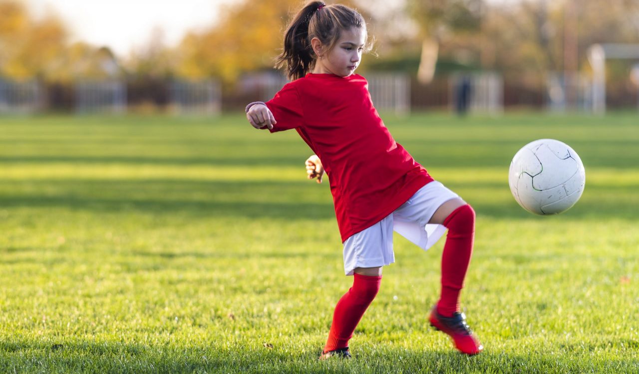 A child wearing a red soccer jersey leans back as she kicks a soccer ball.