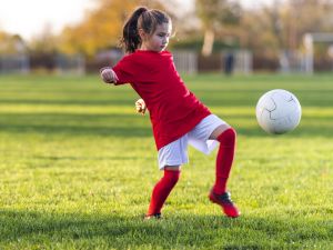 A child wearing a red soccer jersey leans back as she kicks a soccer ball.