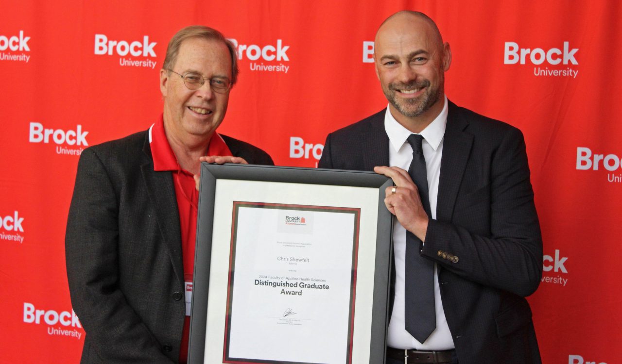 Two men pose in front of a red backdrop holding a framed certificate between them.