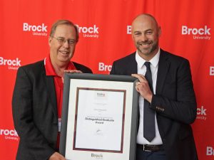 Two men pose in front of a red backdrop holding a framed certificate between them.