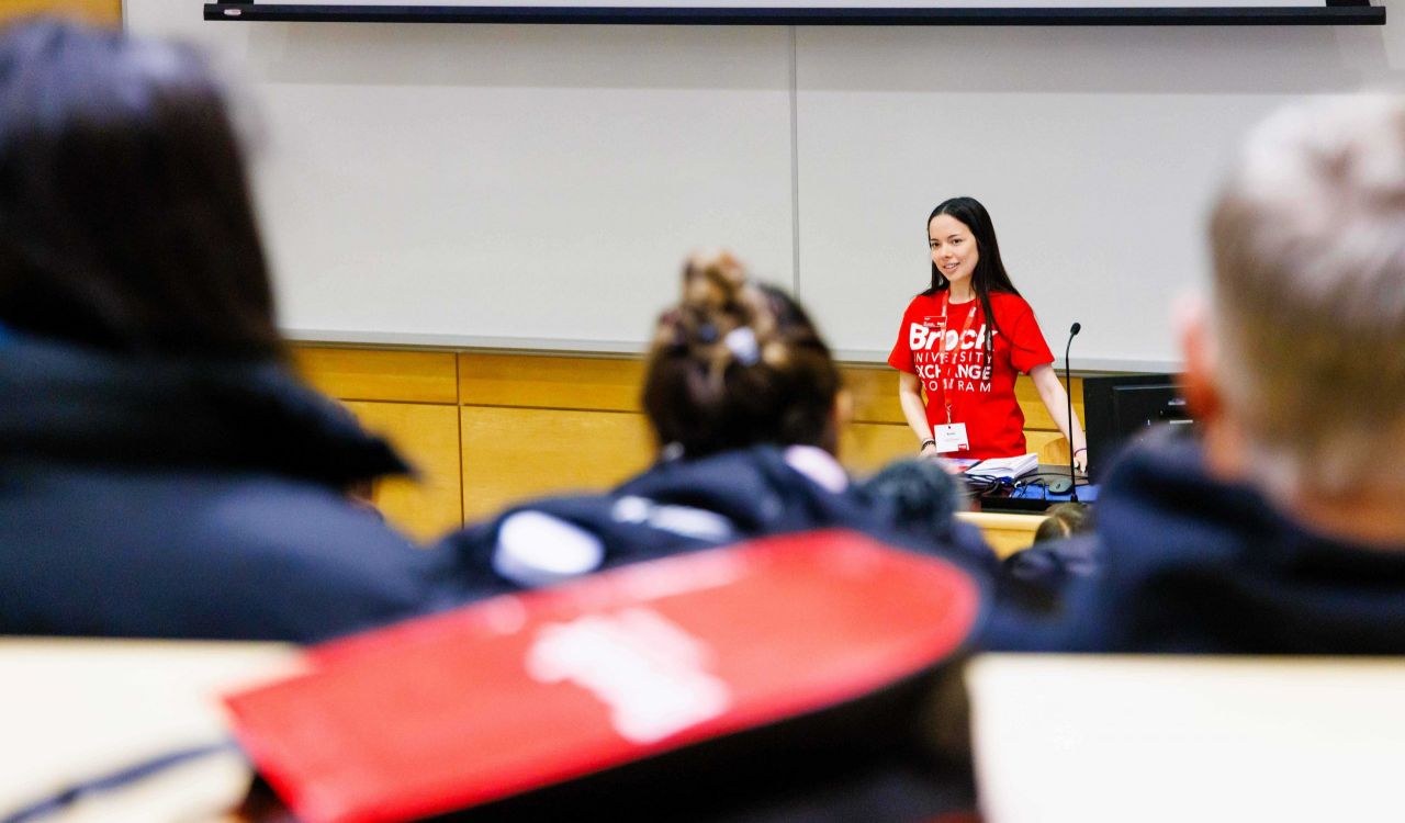 A woman wearing a red Brock University T-shirt stands at the head of a brightly classroom speaking to a full audience at Brock University.