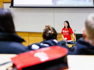 A woman wearing a red Brock University T-shirt stands at the head of a brightly classroom speaking to a full audience at Brock University.