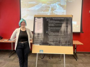 A woman stands in a room with a red wall beside a large poster with text. The woman has dyed bright blue hair and smiles warmly.