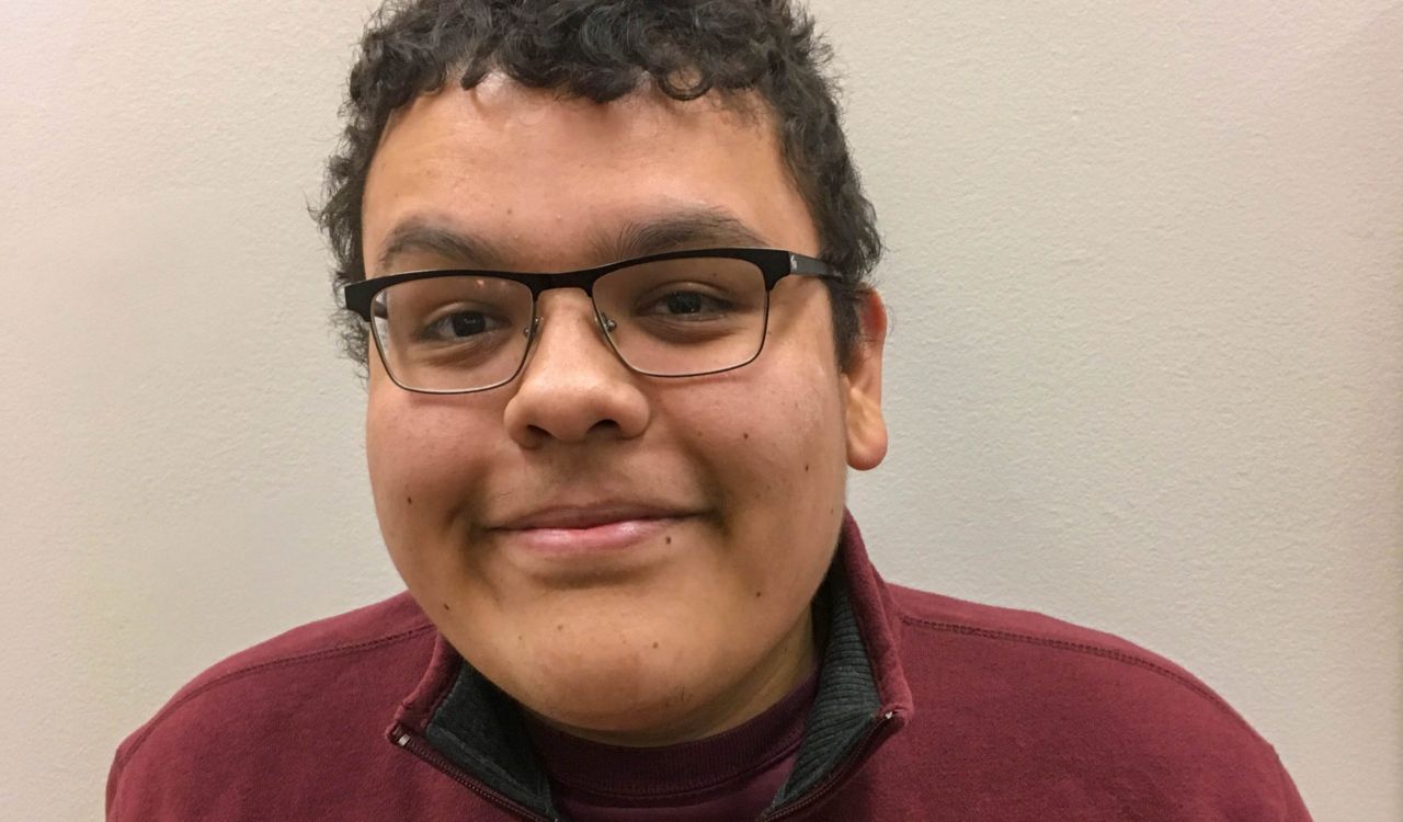 A headshot of Colton Clause standing in front of a beige background wearing a maroon half-zip sweater and glasses.