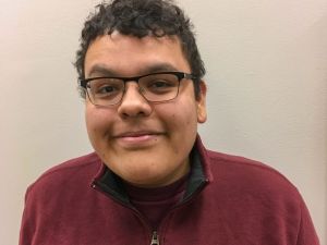 A headshot of Colton Clause standing in front of a beige background wearing a maroon half-zip sweater and glasses.