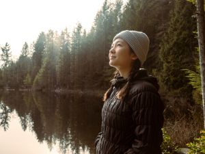 Woman wearing a winter hat and coat stands in front of a lake with trees and looks to the sky.