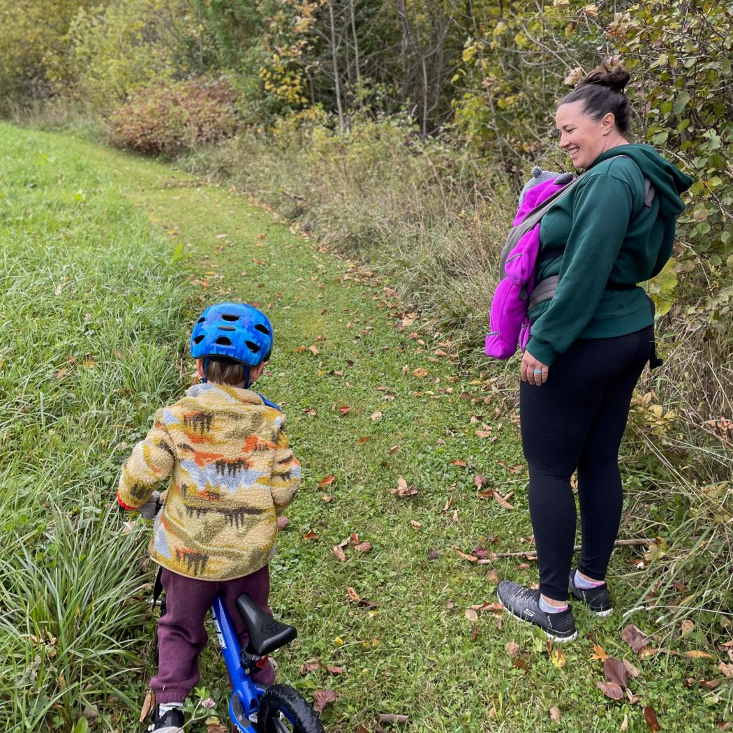 Corliss Bean walking on a trail of mowed grass next to a forest. She carries her infant child in a baby carrier and watches her son ride his bicycle next to her.