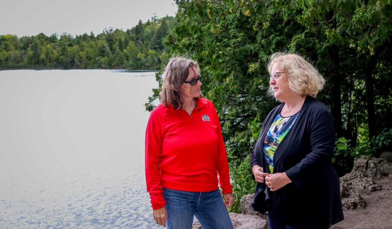 Two women have a conversation beside a lake.