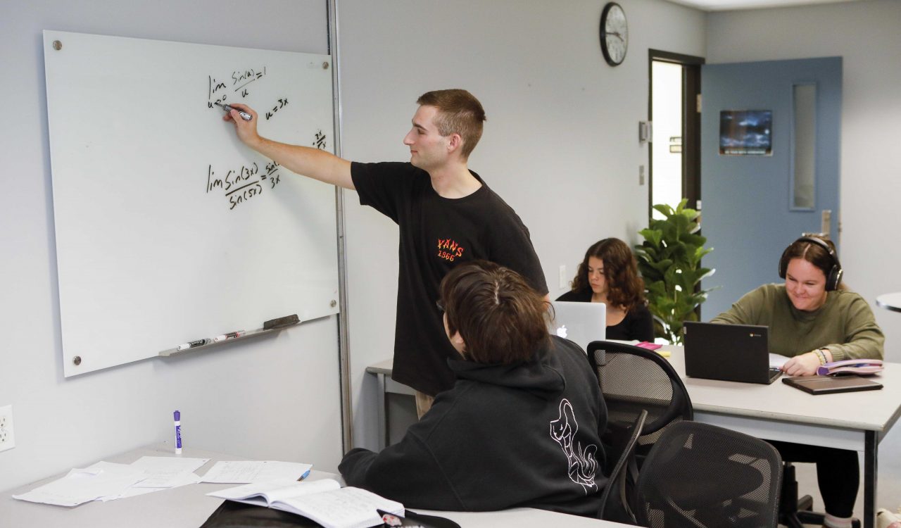 Two students work together to solve a mathematical problem on a whiteboard. Behind them in the background are two other students working independently on laptops.