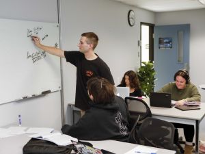 Two students work together to solve a mathematical problem on a whiteboard. Behind them in the background are two other students working independently on laptops.