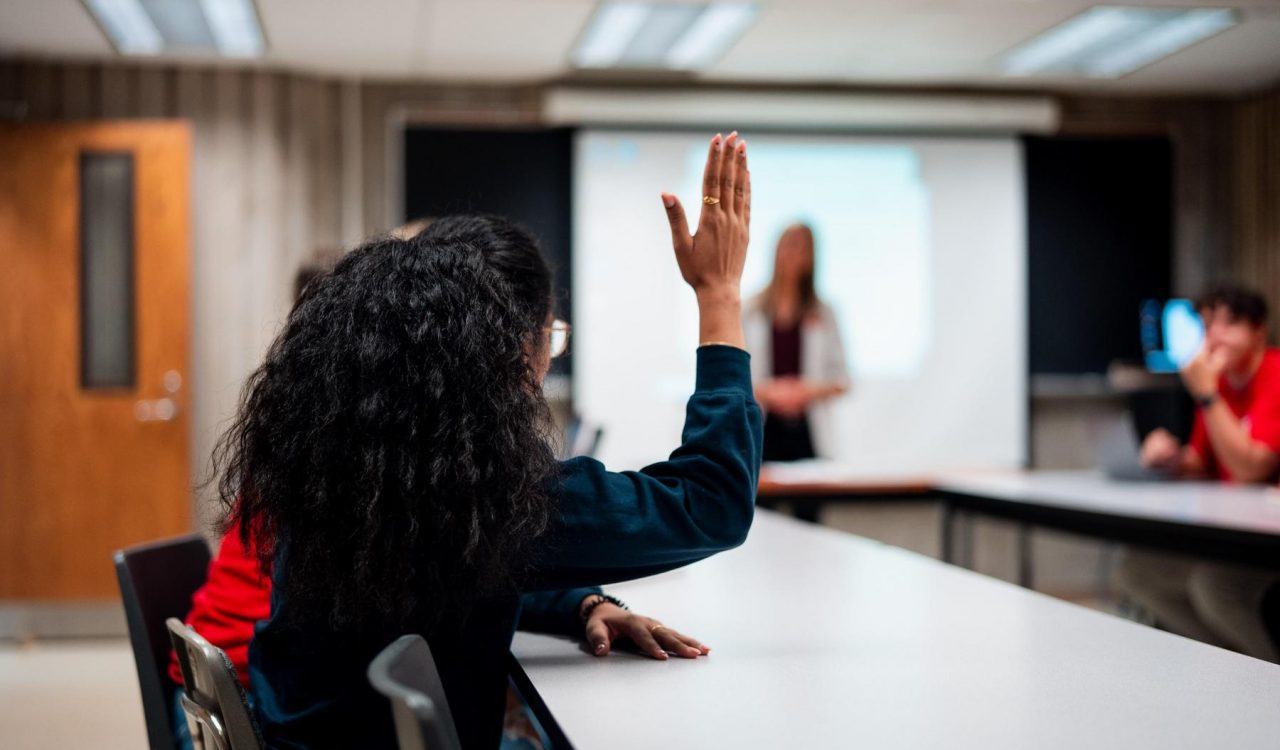 A university student raises a hand while sitting at a table in a small class.