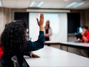 A university student raises a hand while sitting at a table in a small class.