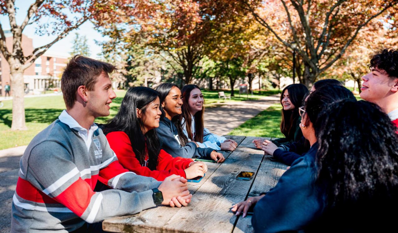 A group of university students sits at a picnic table in a courtyard with fall foliage in the background.