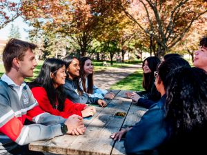 A group of university students sits at a picnic table in a courtyard with fall foliage in the background.