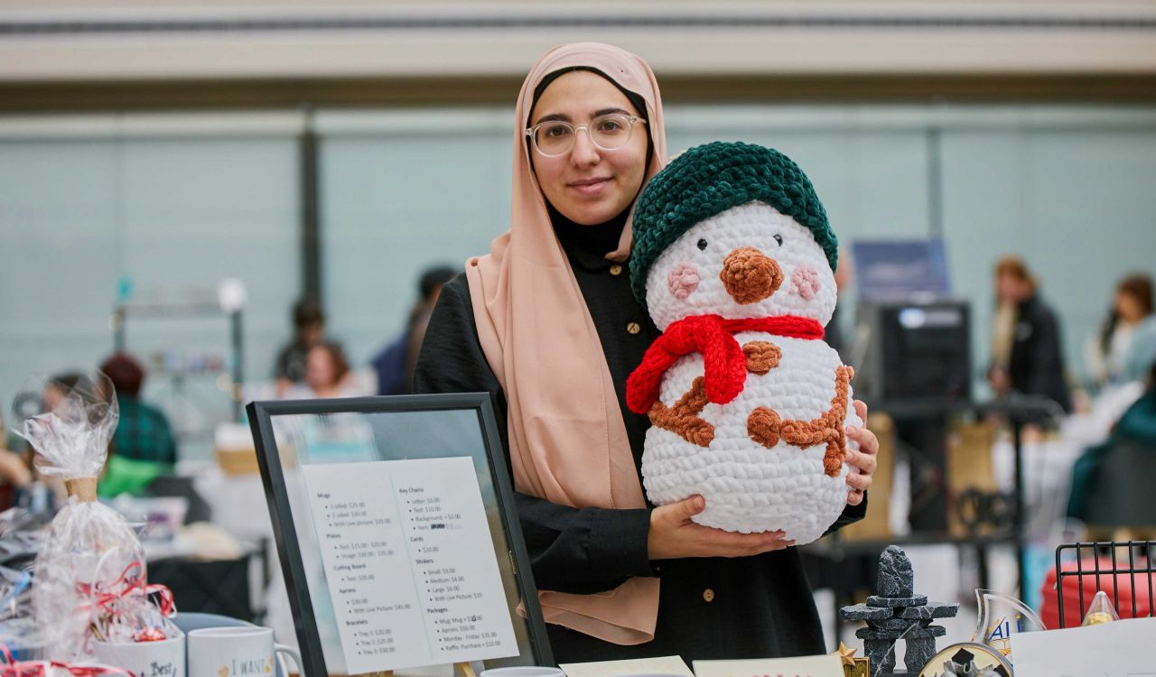 A woman wearing a head covering holds up a stuffed snowman toy at a craft fair.