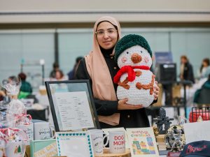 A woman wearing a head covering holds up a stuffed snowman toy at a craft fair.