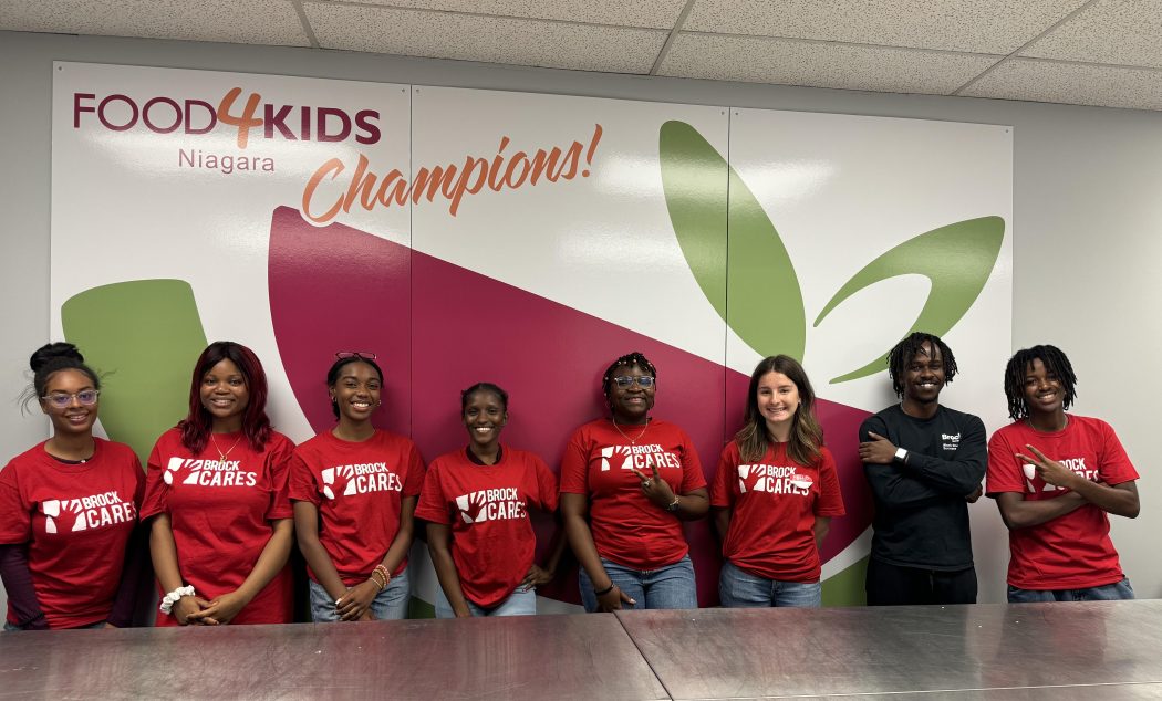 A group of students in red Brock University shirts pose for a photo in a meeting room with the Food 4 Kids Niagara logo on the wall behind them.