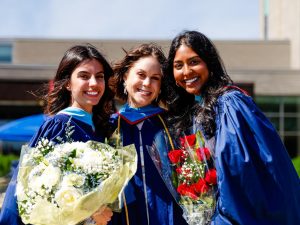 Three women in graduation gowns stand together, two of them holding bouquets of flowers.