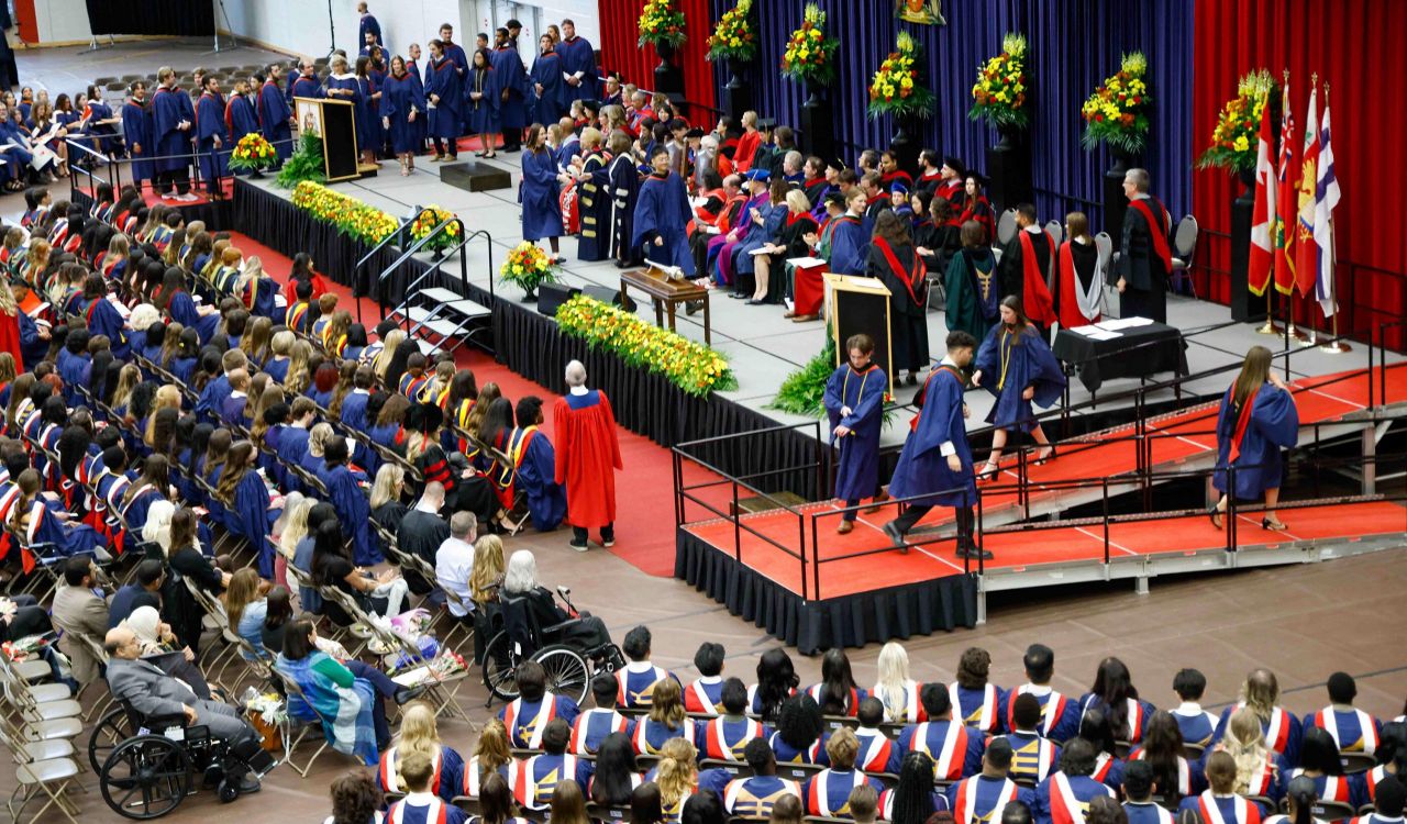 Graduates cross the stage at a graduation ceremony as fellow grads look on in an auditorium.