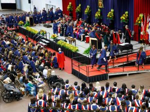 Graduates cross the stage at a graduation ceremony as fellow grads look on in an auditorium.