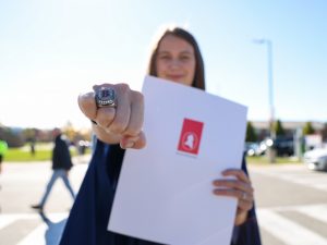 A graduate holds out a ring with a large B while holding her degree.