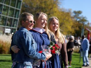 A graduate in a blue gown holds flowers while being hugged by two women.