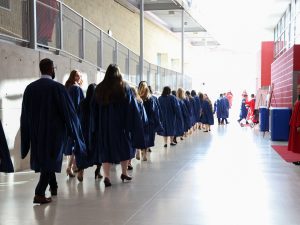 A lineup of graduates in blue gowns walk down a hallway.