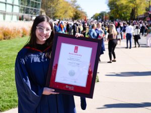 A graduate holds a framed degree while standing outside with a crowd in the background.