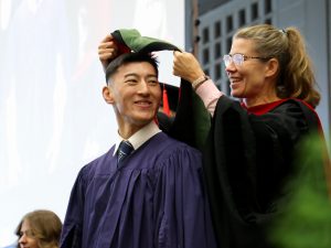 A woman places a hood over the head of a graduate on stage.