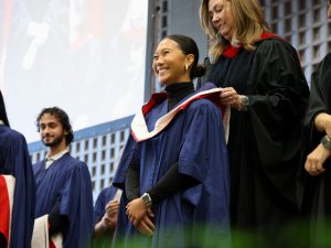 A graduate smiles while a hood is placed around her next on stage at Convocation.