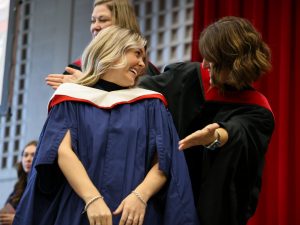 A graduate looks backwards smiling at the person who has just placed a hood on her shoulders.