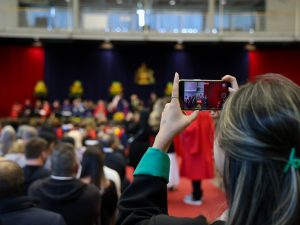 A woman stands at the back of an auditorium filming a graduating on her phone.