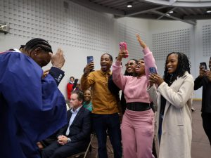 A graduate is celebrated by a group of people who cheer and take photos with their phones.