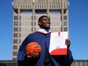 A man in a graduation gown holds a basketball in one hand and his degree in the other.