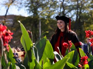 A woman in a graduation gown walks outdoors with flowers in the foreground.