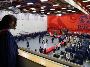 A graduate looks over the ledge of a balcony, where graduates are getting their gowns on below.