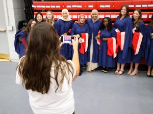 A group of graduates stand together having their photo taken by a woman holding a phone.