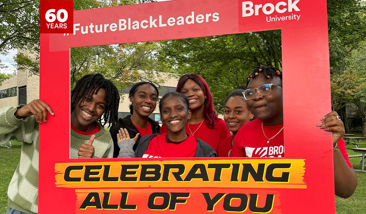 A group of high school and first-year university students pose for a photo outside holding a large red frame.