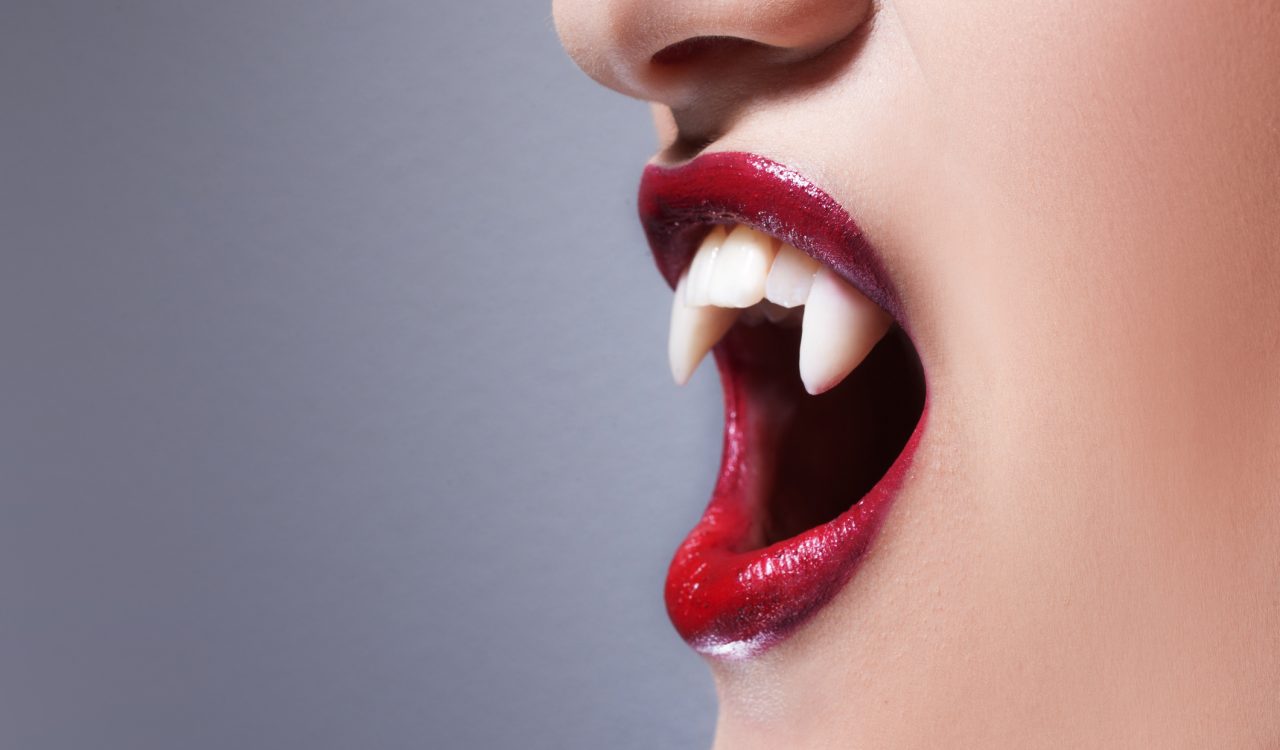 Close-up of mouth of a woman a woman wearing fake vampire teeth and red lipstick.