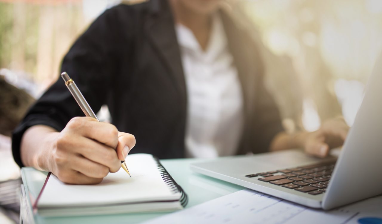 Close-up of a person's hand writing on a notebook beside a laptop.