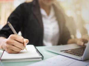 Close-up of a person's hand writing on a notebook beside a laptop.