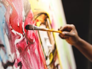 A hand holds a paint brush as it applies brown paint to a colourful abstract painting.