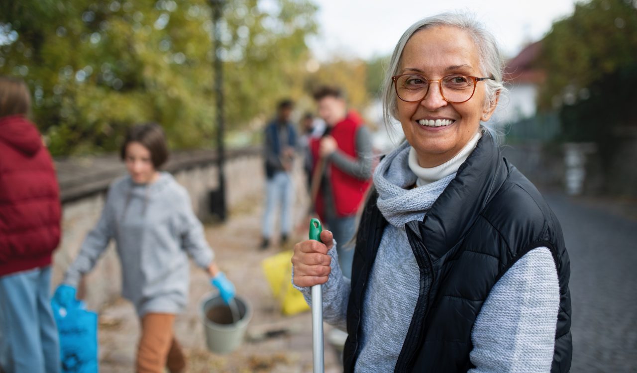 Close-up of older woman volunteer smiling into the camera, gripping the top of a rake, with other volunteers cleaning up the environment fuzzed out in the background.