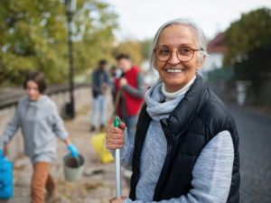 Close-up of older woman volunteer smiling into the camera, gripping the top of a rake, with other volunteers cleaning up the environment fuzzed out in the background.