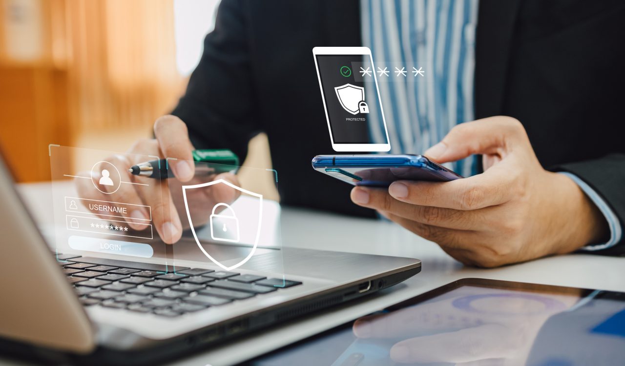 Close-up view of a person's hands holding a smartphone with cybersecurity icons floating above it. The person is also holding a pen and is seated at a desk with a laptop on it.