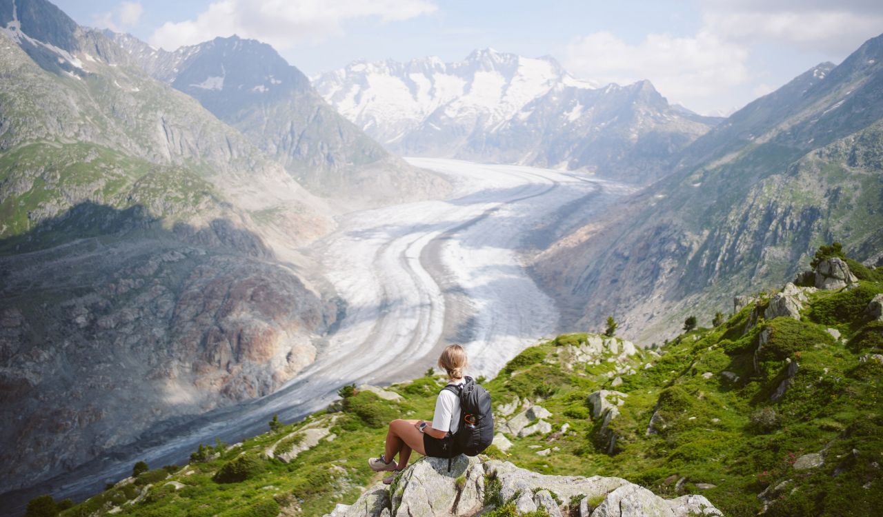 Woman with blonde hair wears a t-shirt and sits at the base of a snow- and ice-covered mountain with green grass showing through.