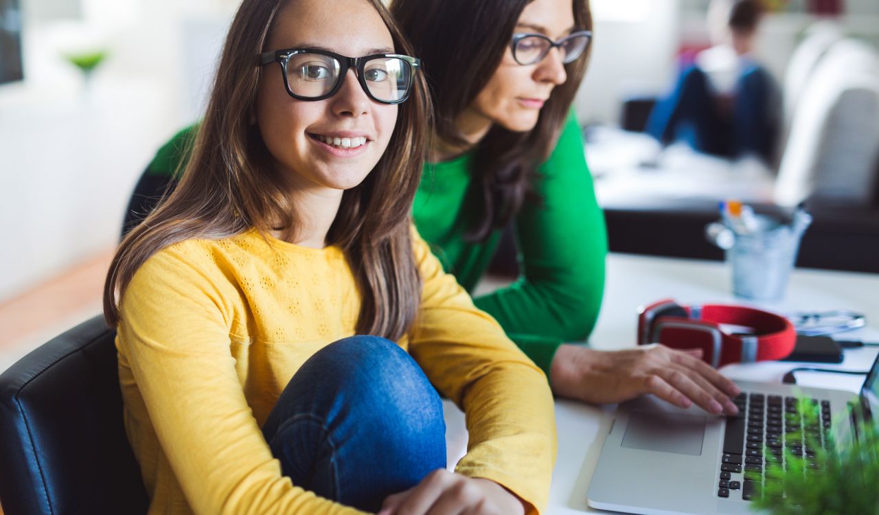 A girl wearing glasses and a yellow sweater sits next to a woman operating a computer at a desk in a naturally lit room.