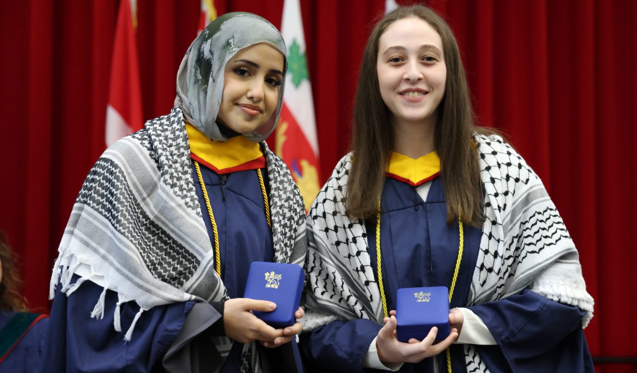 Two women in graduation gowns stand together, each holding a blue box containing a medal.